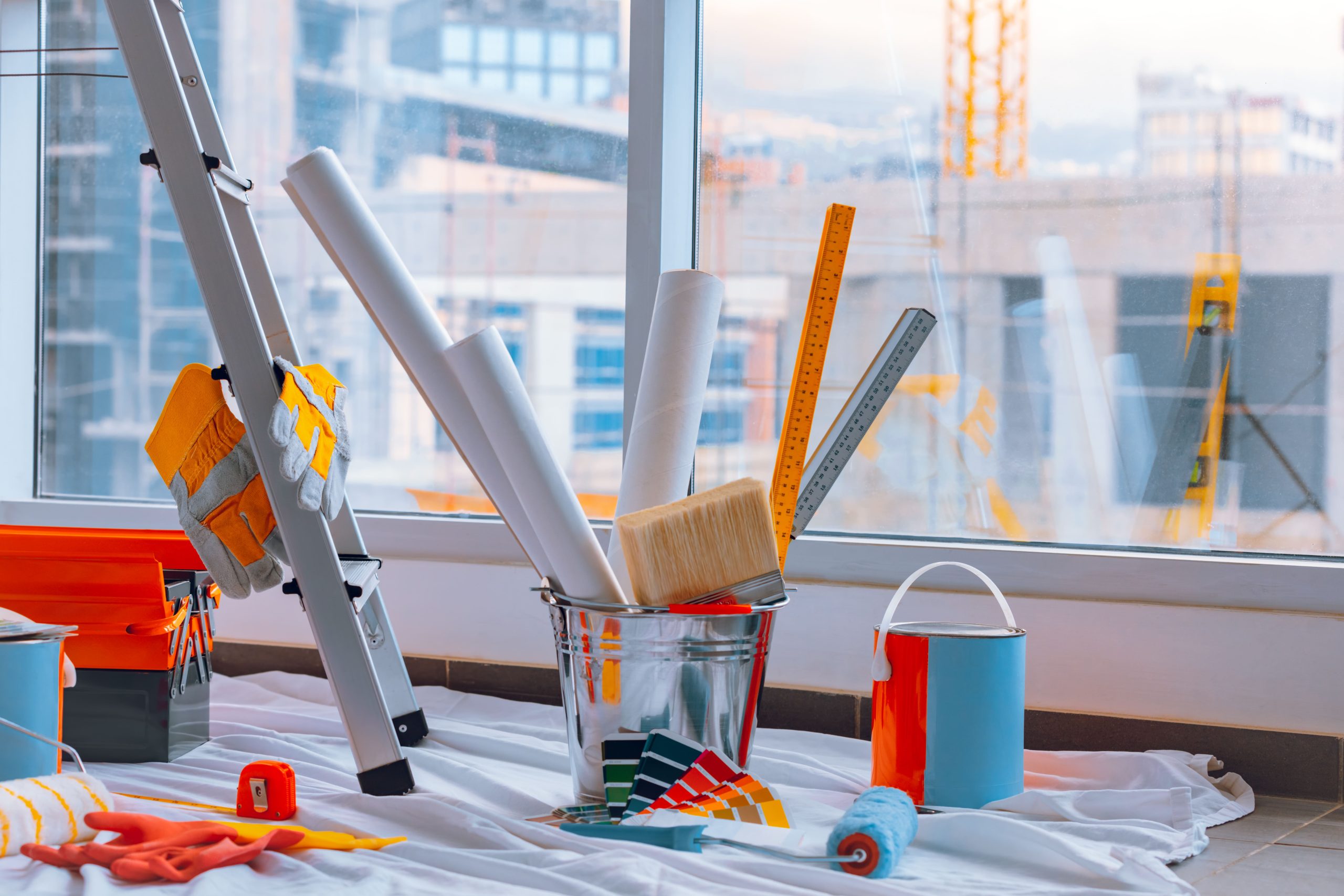 Paint and renovation tools including paintbrush, rollers, color swatches, gloves, ladder, blueprints, rulers, and paint cans near a window.