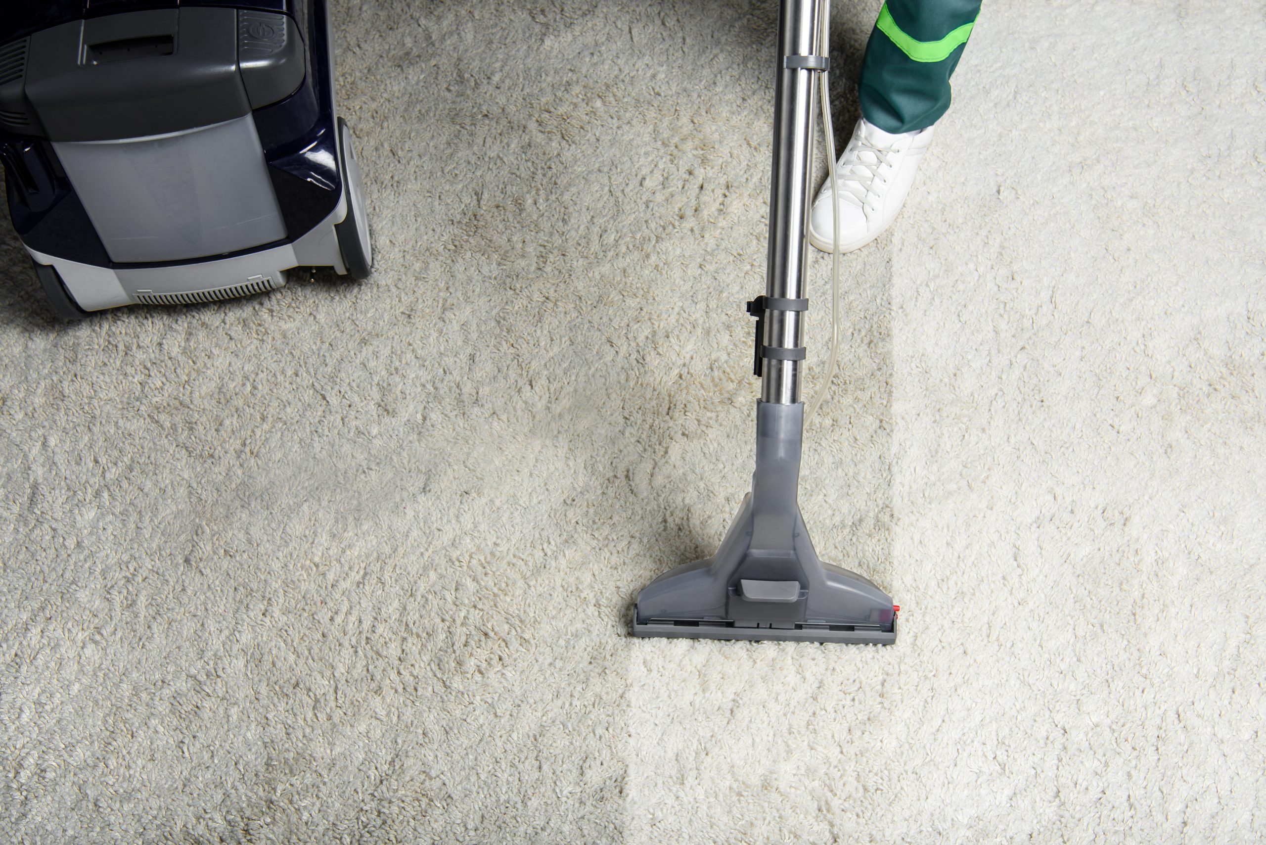 Vacuum cleaner removes dirt from a light carpet, leaving a visibly clean path; a person in sneakers operates the floor-cleaning machine.