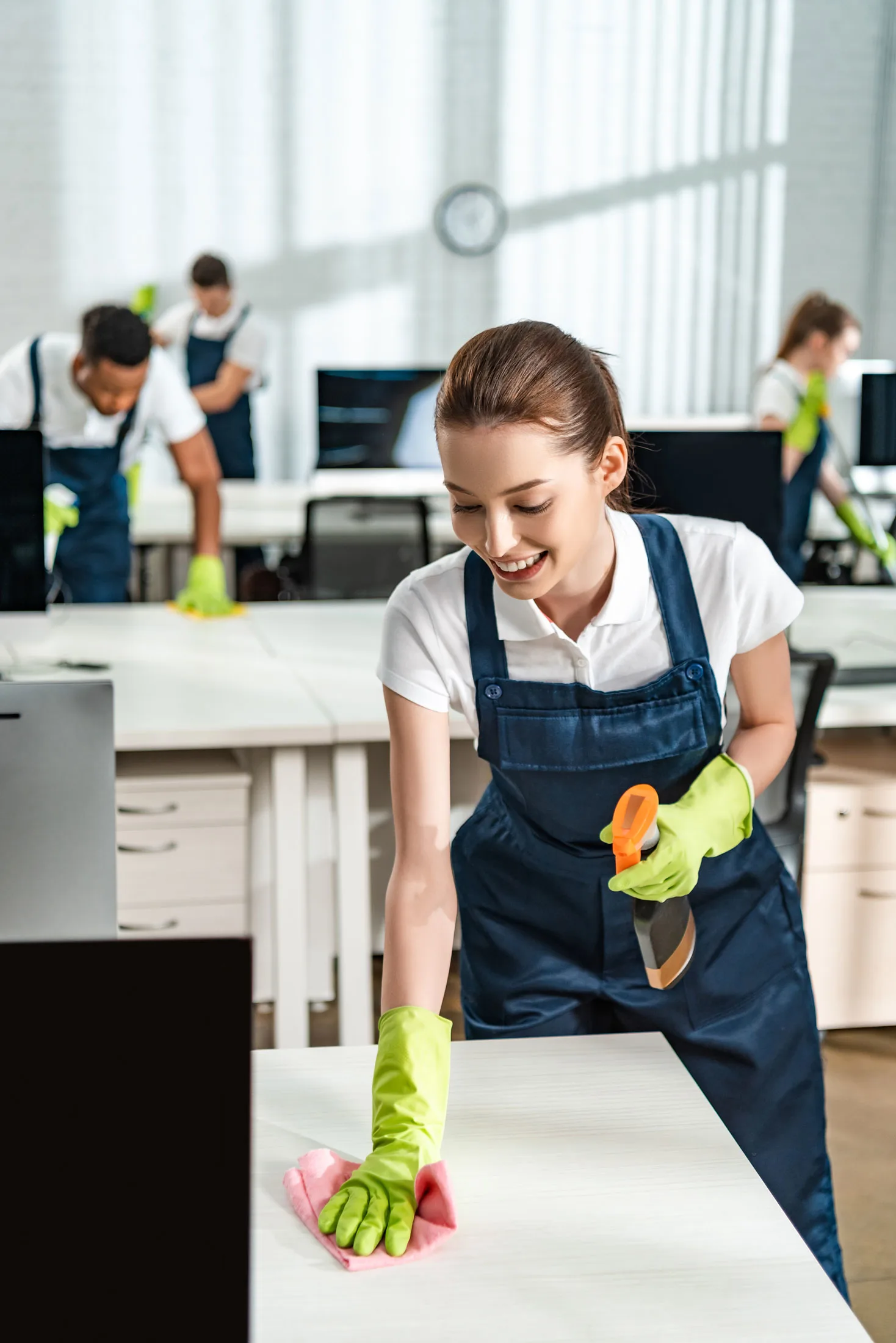 A smiling woman in green gloves cleans a desk with a cloth, while others work in the background. The setting appears to be a modern office environment.