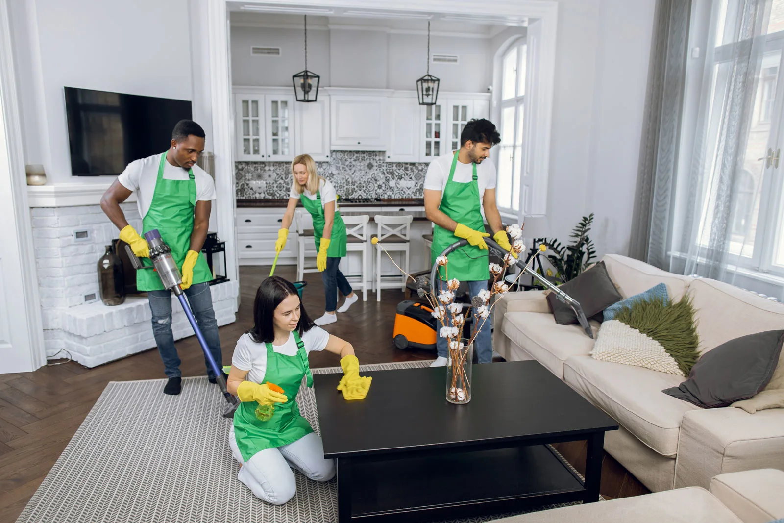 A team of four cleaners in green aprons is actively cleaning a modern living room, using various cleaning tools. The space features a light-colored sofa, a coffee table, and a stylish kitchen area in the background.