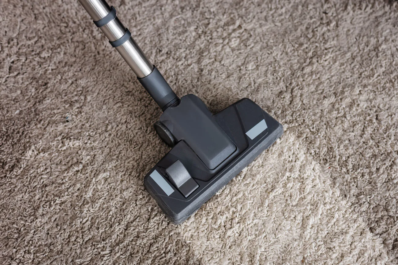A vacuum cleaner head is shown on a carpet, highlighting a cleaned area next to an uncleaned section. The image emphasizes the effectiveness of a cleaning service.